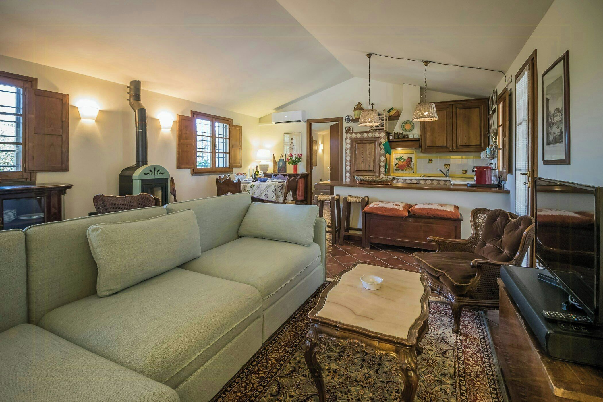 A living room with a sofa, coffee table, and a fireplace. There is a dining area with a table and chairs. The kitchen is visible with cabinets and a hanging light.