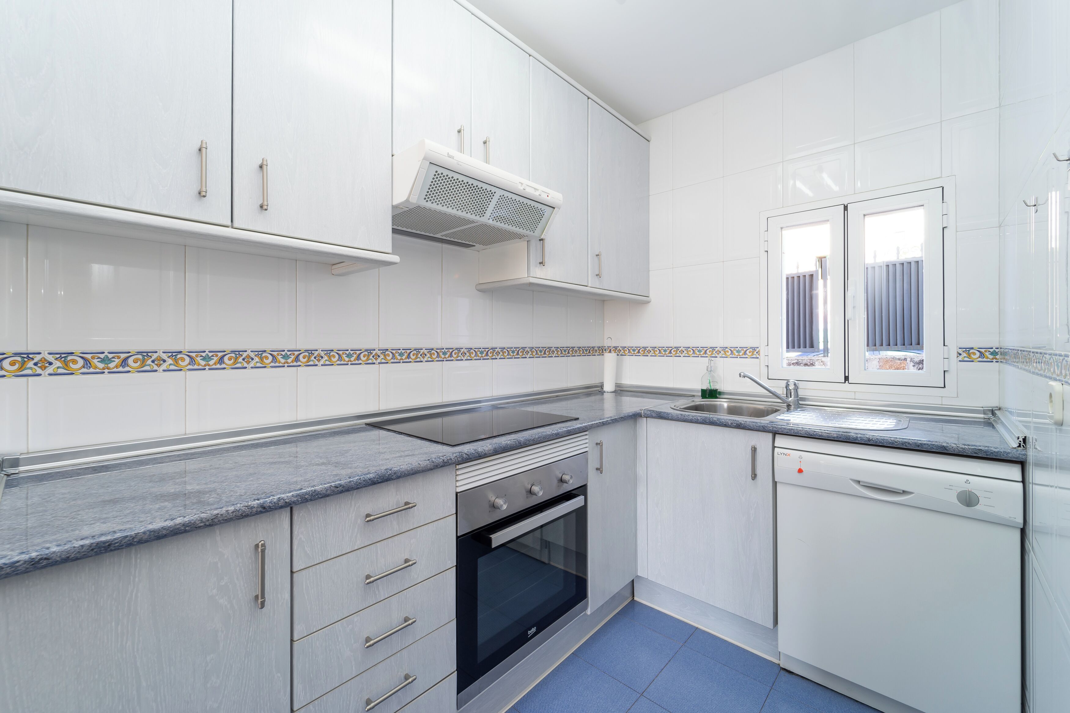 A modern kitchen with white cabinets, a built-in oven, a dishwasher, and a window with blinds.