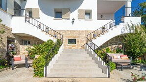 A white building with a stone facade, a staircase with black railings, and outdoor seating with cushions.