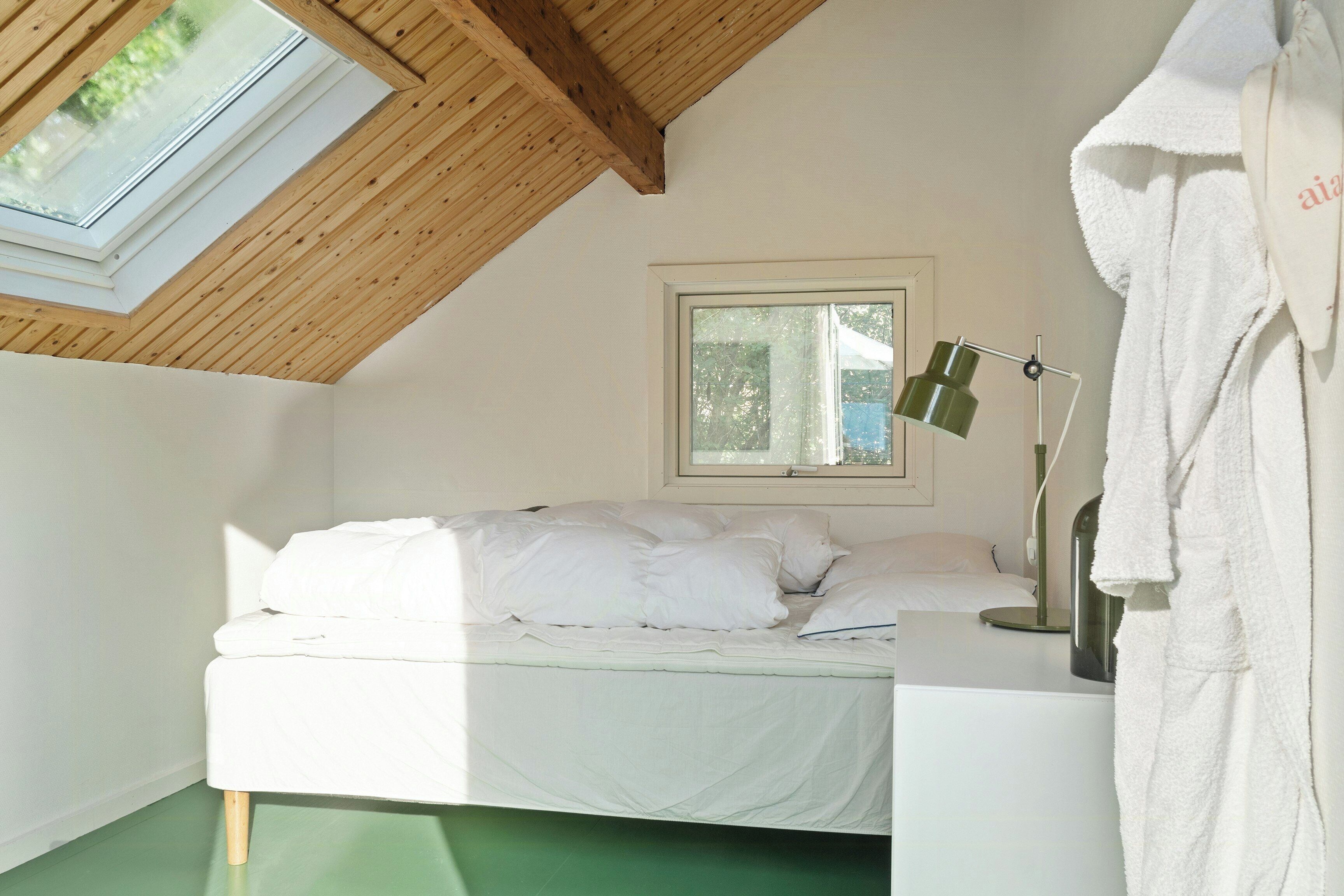 Quarto com teto inclinado, uma cama com roupa de cama branca, luminária de cabeceira e uma janela.