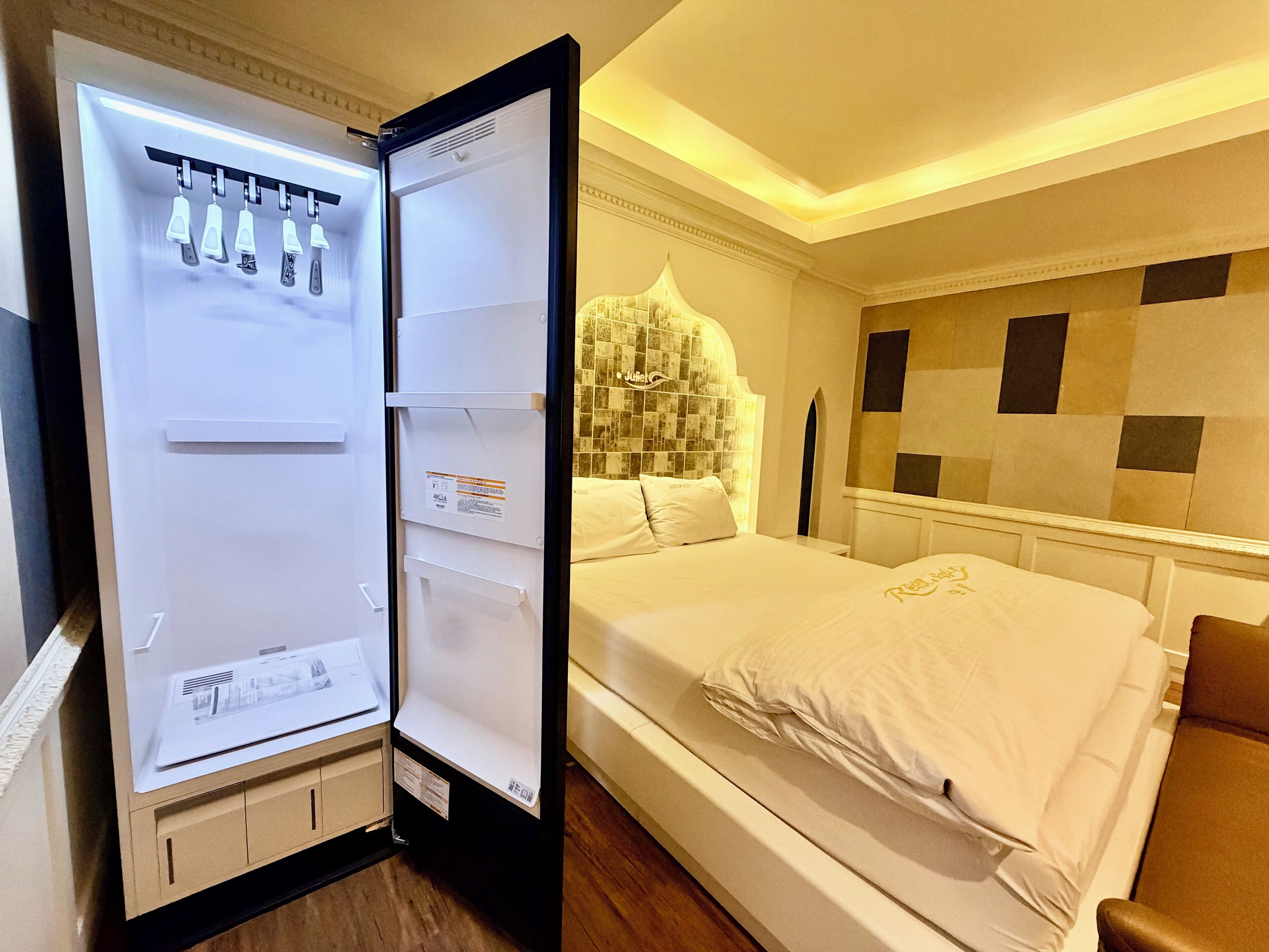 A hotel room with a bed, a white headboard, a wall with a checkered pattern, and a refrigerator with a control panel.