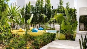 Um jardim com degraus que levam a uma piscina, rodeado por diversas plantas e um edifício branco ao fundo.