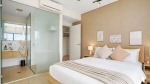 Moderni hotellihuone, jossa on lasiseinäinen suihkukaappi, puurunkoon rakennettu sänky, yöpöytä lampun kanssa ja kattotuuletin.