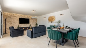 A modern living room with a stone wall, a flat-screen TV, a sofa, a dining table with chairs, and a staircase. - Villa Flora Zadar Villas (Benkovac)