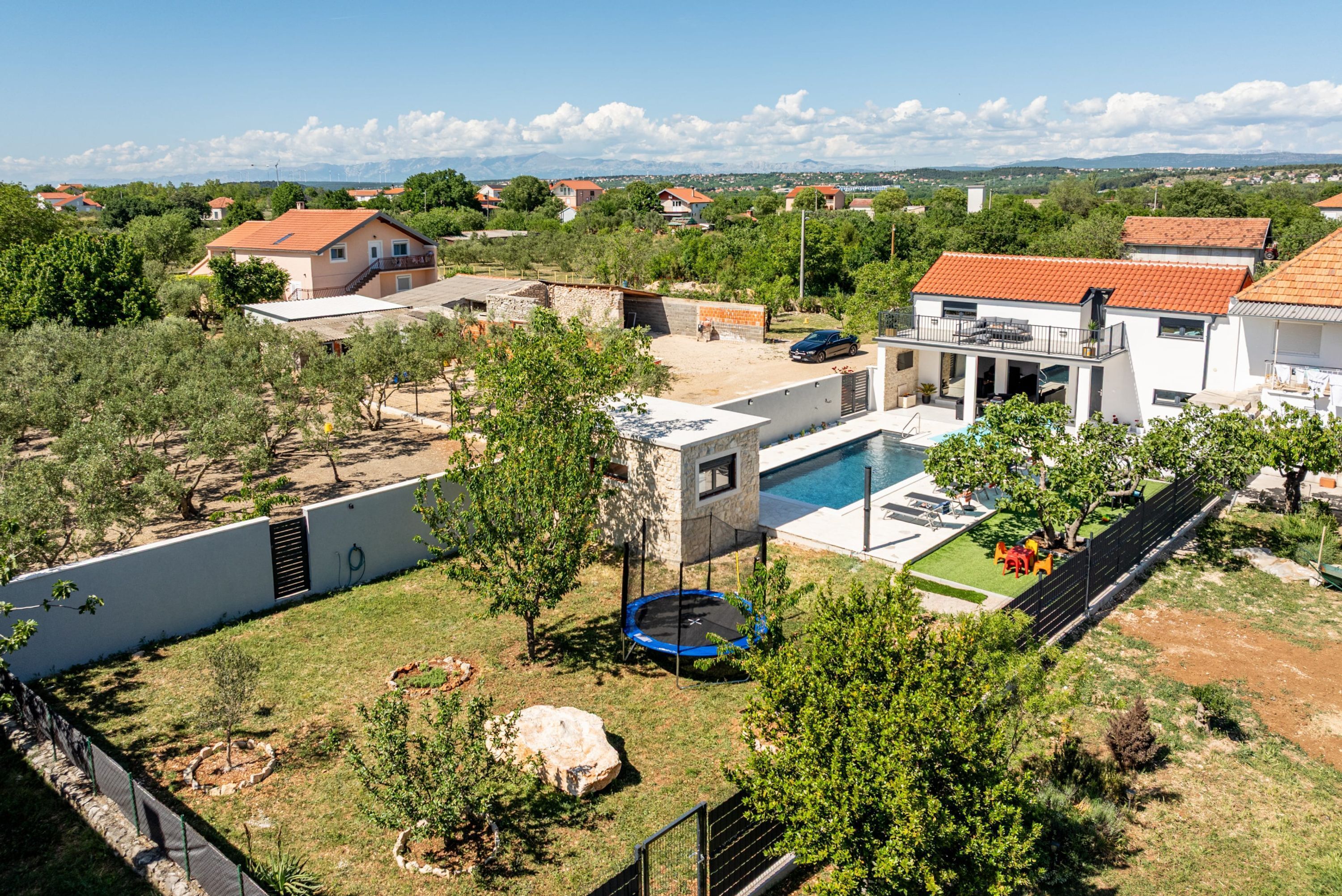 A residential area with a swimming pool, a trampoline, and a view of distant houses and mountains.