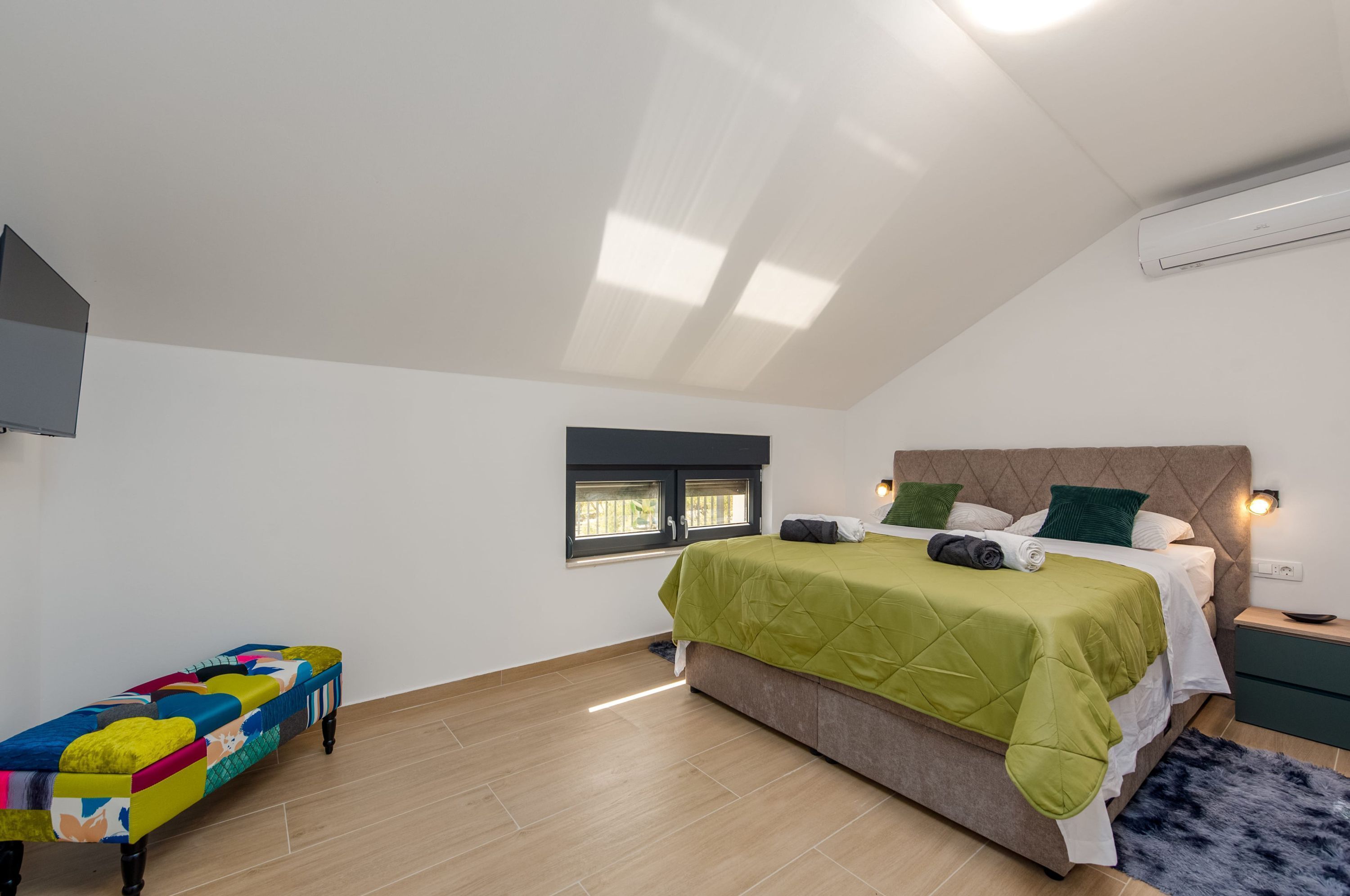 Een slaapkamer met een bed, een bankje, een raam en een airconditioning.