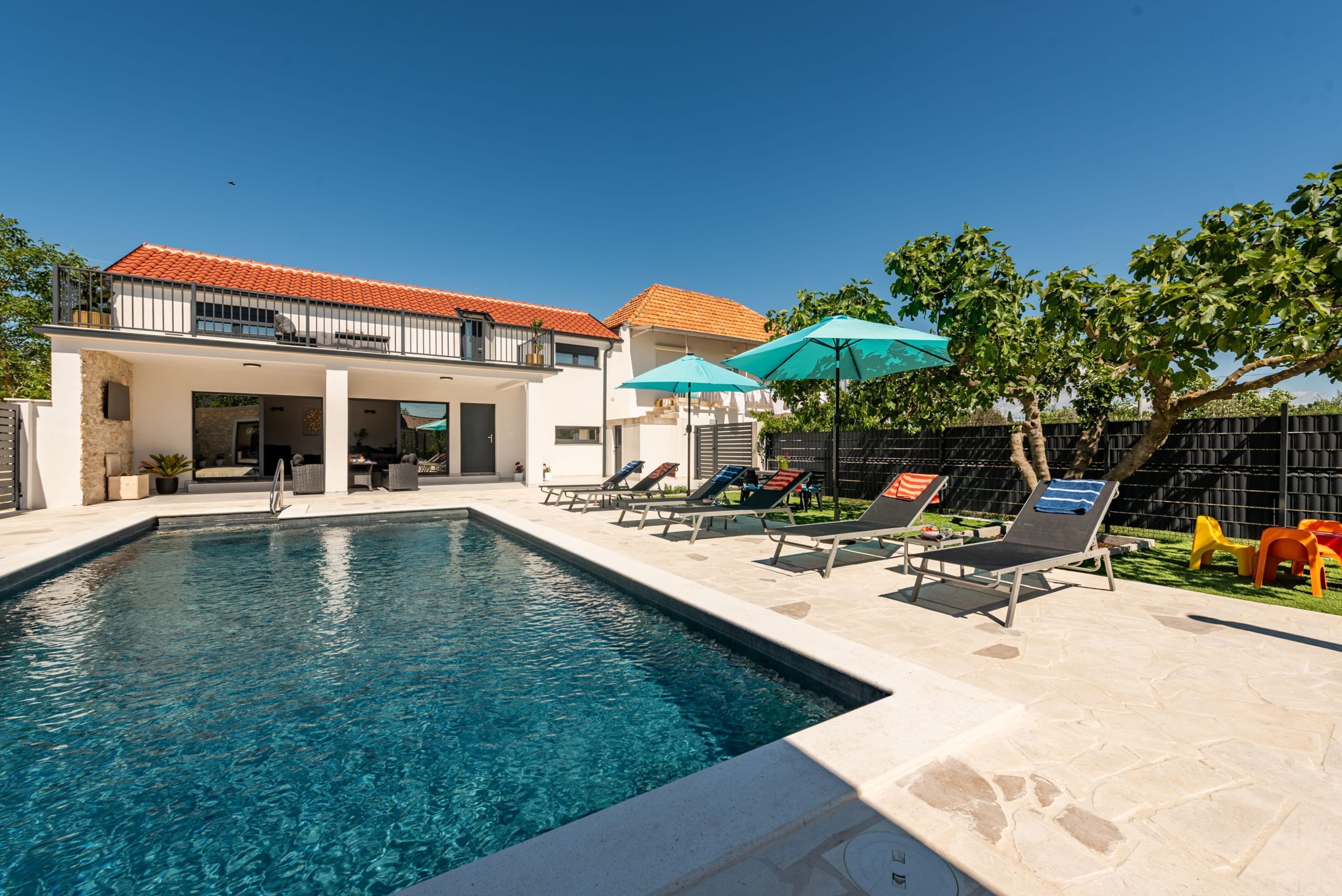 A swimming pool with lounge chairs and umbrellas, a building with a red-tiled roof, and a fenced yard.