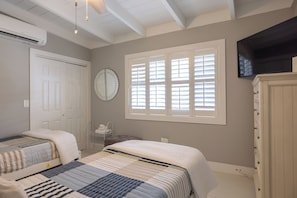A bedroom with two beds, a large window with blinds, a clock on the wall, and a TV mounted above a cabinet. - Unit 196 - 500 4th Street: 2  BR, 2  BA Duplex in Key Colony Beach, Sleeps 4 (Key Colony Beach)