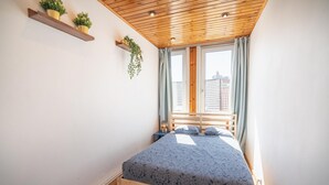 Quarto com teto de madeira, cama com colcha estampada, janela com cortinas e prateleira com plantas.