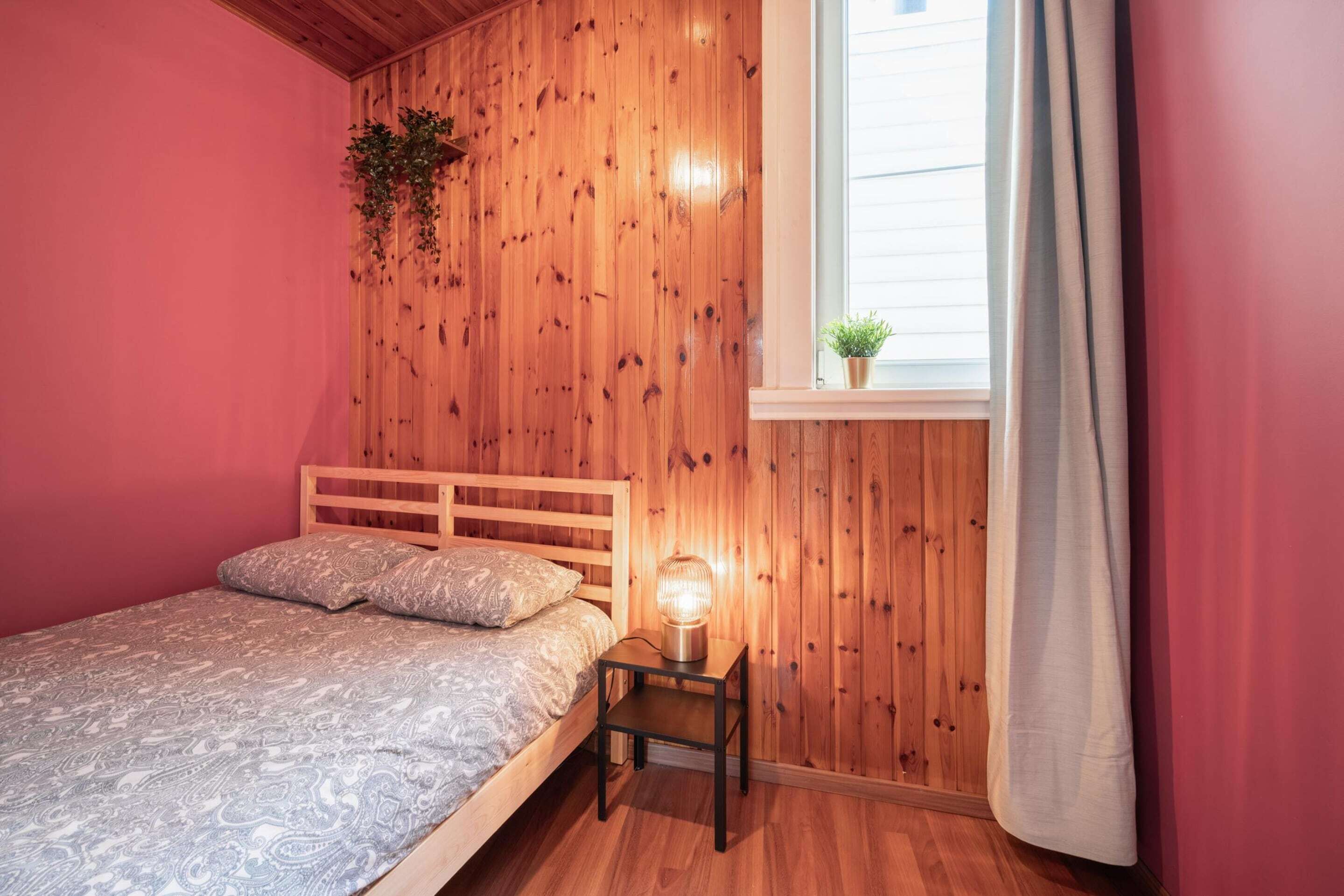 A bedroom with a wooden headboard, a bedside table with a lamp, a window with a curtain, and a plant on the wall.