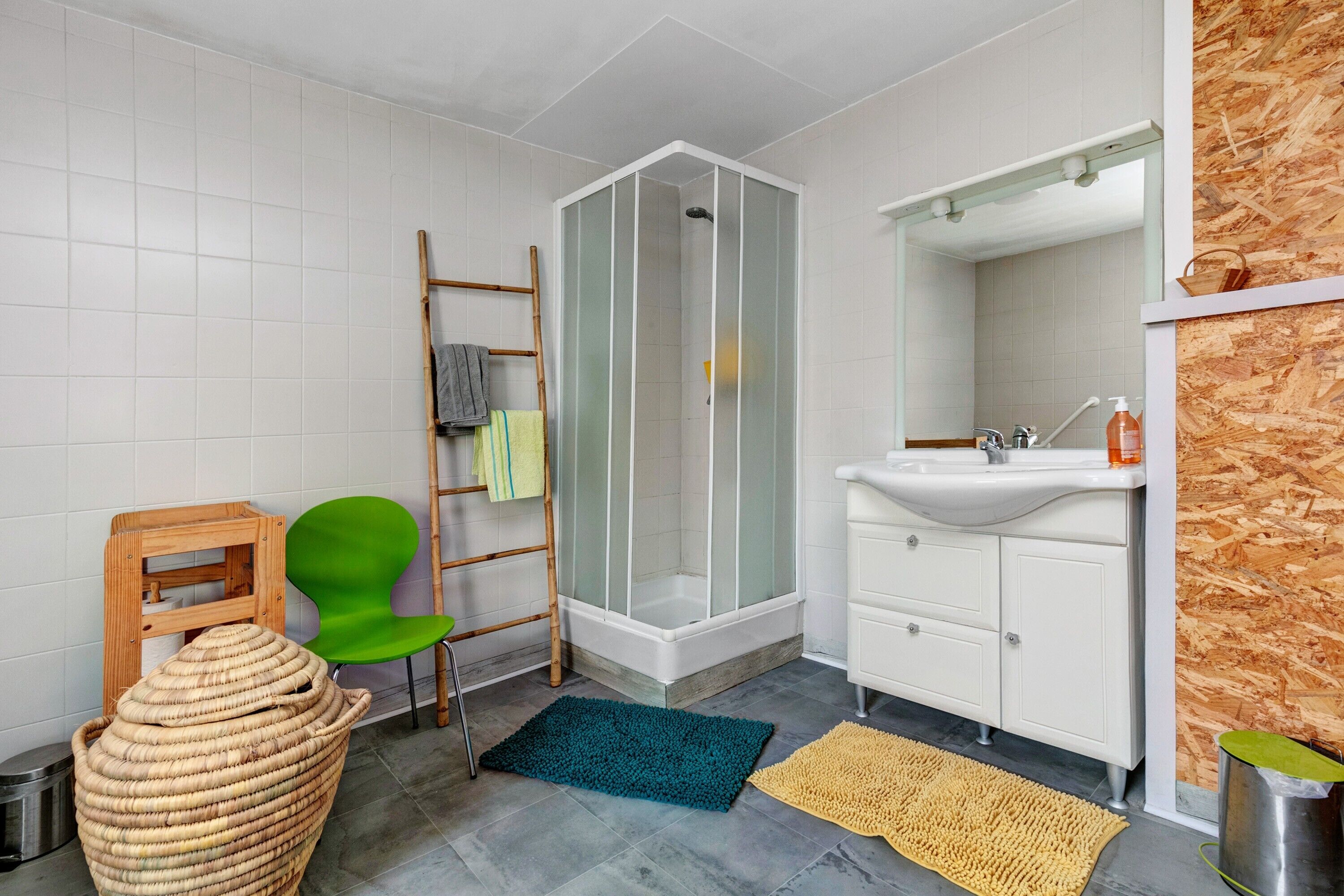 Een badkamer met een douche, een wastafel, een spiegel, een groene stoel, een rieten mand en een houten wand.