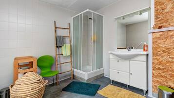 Een badkamer met een douche, een wastafel, een spiegel, een groene stoel, een rieten mand en een houten wand.