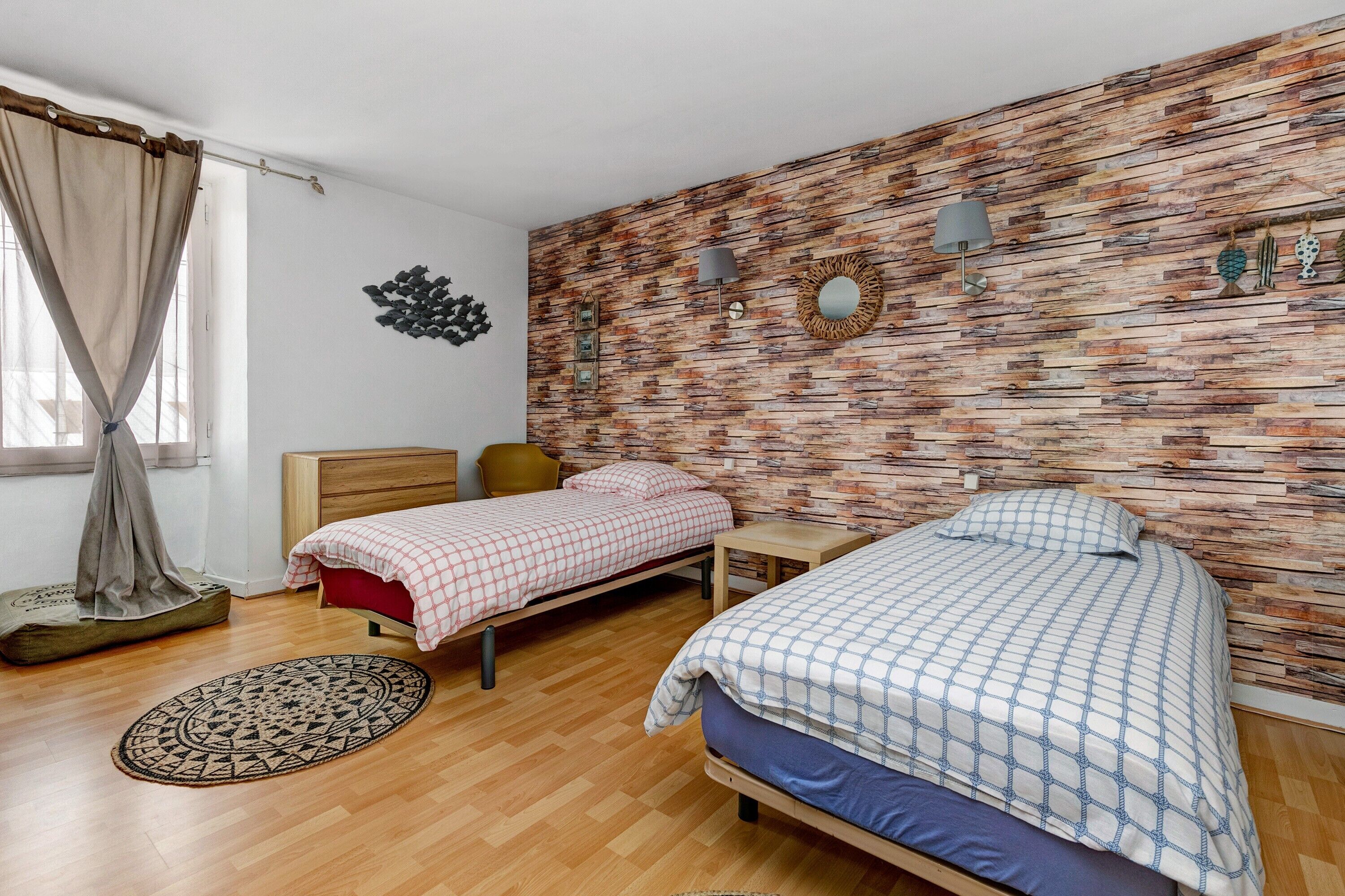A bedroom with two beds, a wooden floor, a brick-patterned wall, a window with curtains, and a wall-mounted lamp.