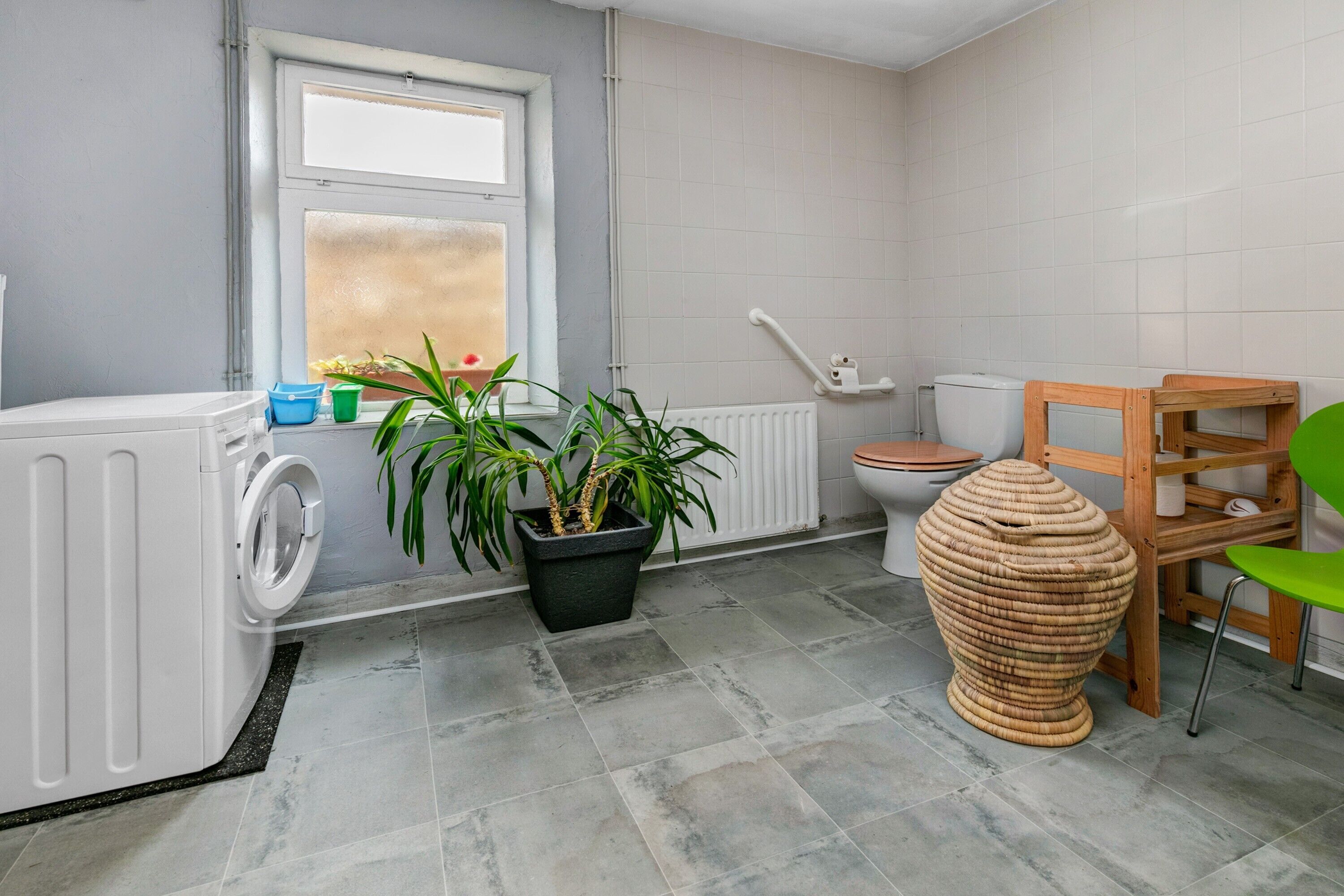 Kylpyhuoneessa on pesukone, patteri, wc-istuin, ruukussa oleva kasvi ja punottu tuoli.