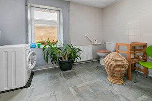A bathroom with a washing machine, a radiator, a toilet, a potted plant, and a wicker chair.