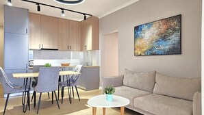 A modern kitchen with wooden cabinets, a dining table with chairs, a round white table with a plant, and a beige sofa. - Deep Blue Rooftop - 111 (Sarandë)