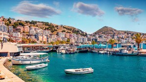 A marina with boats docked, a waterfront promenade, and a hillside town in the background. - Deep Blue Rooftop - 111 (Sarandë)