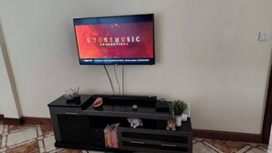A flat-screen TV mounted on a wall above a black entertainment center with a glass bowl, a teddy bear, and a small plant. - Spacious 2br, 3BA Great Location (Nairobi)