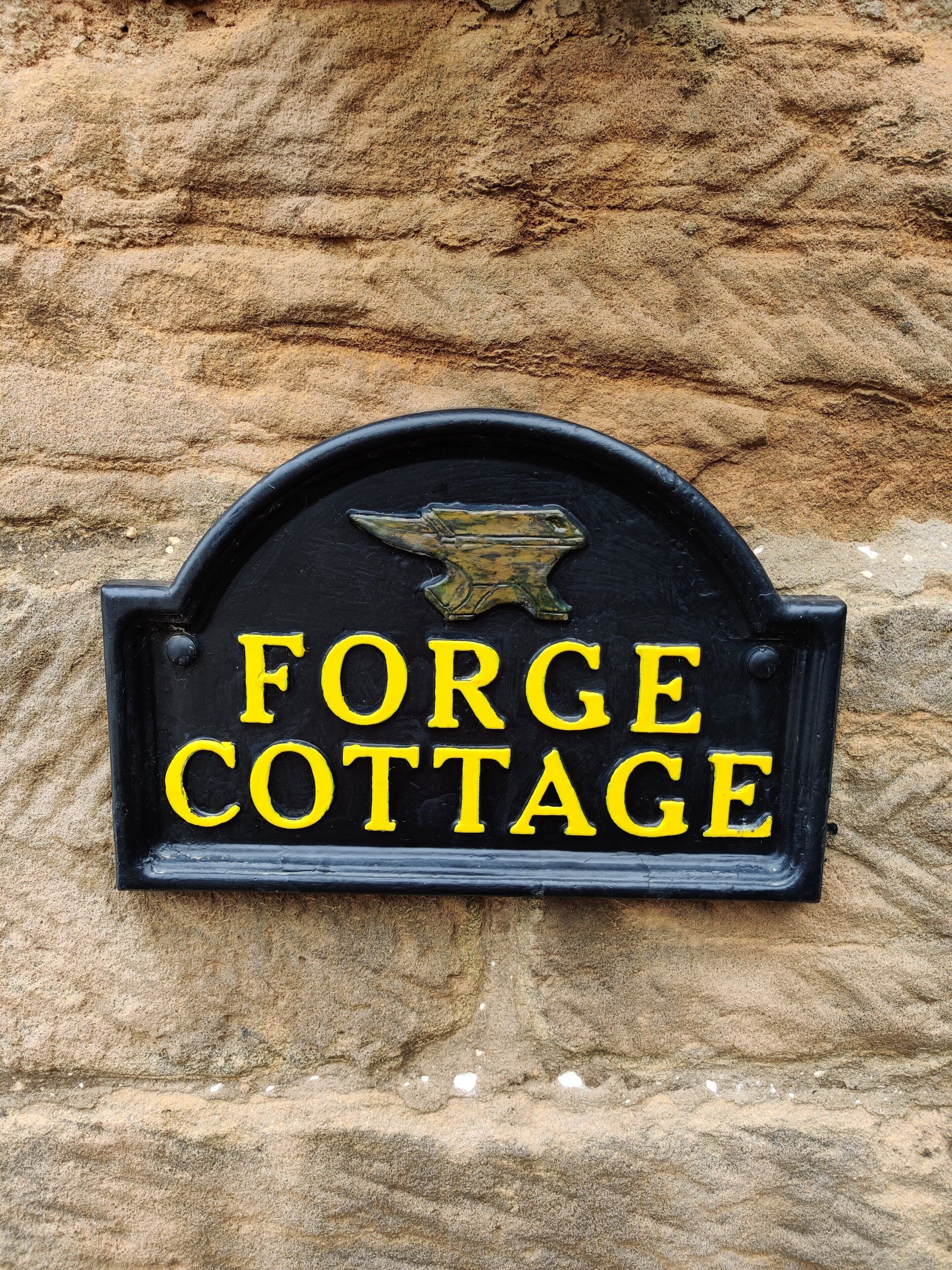 Une plaque en métal noir avec des lettres jaunes, fixée à un mur de pierre, sur laquelle on peut lire : « FORGE COTTAGE ».