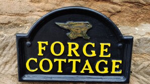 Ein schwarzes Metallschild mit gelber Schrift auf einer Steinmauer, mit der Aufschrift 'FORGE COTTAGE'.