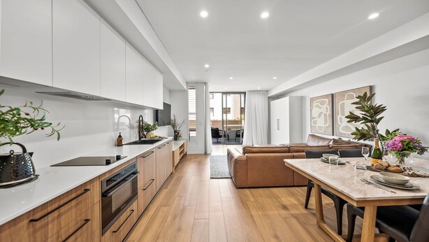 A modern kitchen with white cabinets, a central island, and a dining area with a table and chairs. - Kove Apartment - G01 (Kiama)