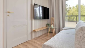 Una habitación moderna con una televisión de pantalla plana montada en la pared, piso de madera, cama con cubrecama blanco, una mesita de vidrio con una planta y una ventana con cortinas.