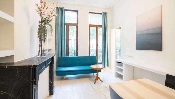Een moderne kamer met een blauwe bank, een klein rond tafeltje, een marmeren aanrecht en een schilderij aan de muur.