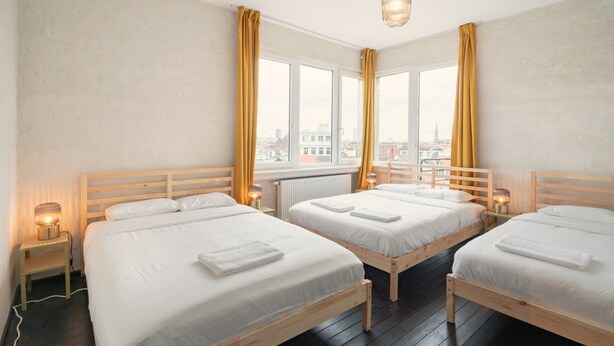 A hotel room with three wooden beds, a large window with curtains, a hanging light fixture, and a bedside table with a lamp. - 2BR Apartment Near Grand Central Station (Antwerp)