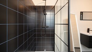 A modern bathroom with dark tiled walls, a glass shower enclosure, and a white sink. - 2BR Apartment Near Grand Central Station (Antwerp)