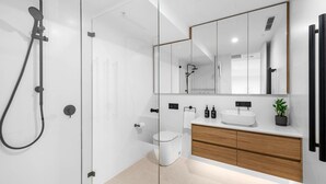 A modern bathroom with a glass shower enclosure, a white toilet, a wooden vanity with a sink, and a large mirror. - Kove Apartment - B01 (Kiama)
