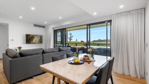 A modern living room with a sofa, a dining table with a vase of flowers, and a large window with a view of a golf course. - Kove Apartment - B01 (Kiama)