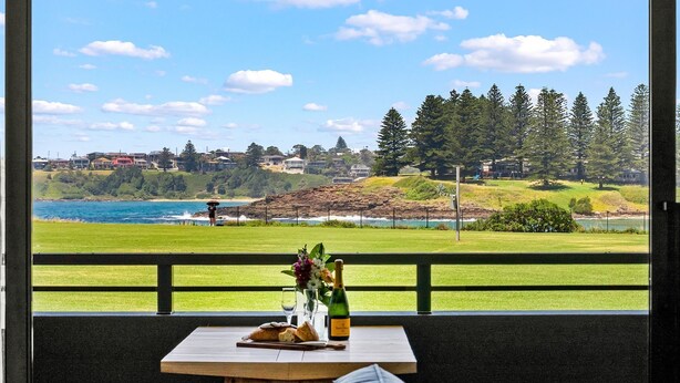 A table with a bottle and glasses, a vase with flowers, and a view of a coastal landscape with houses and trees. - Kove Apartment - B01 (Kiama)