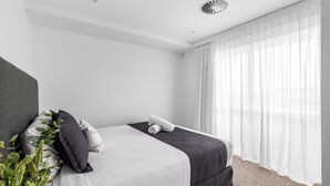 A modern hotel room with a bed, a bedside table, a plant, and sheer curtains. - Kove Apartment - B01 (Kiama)