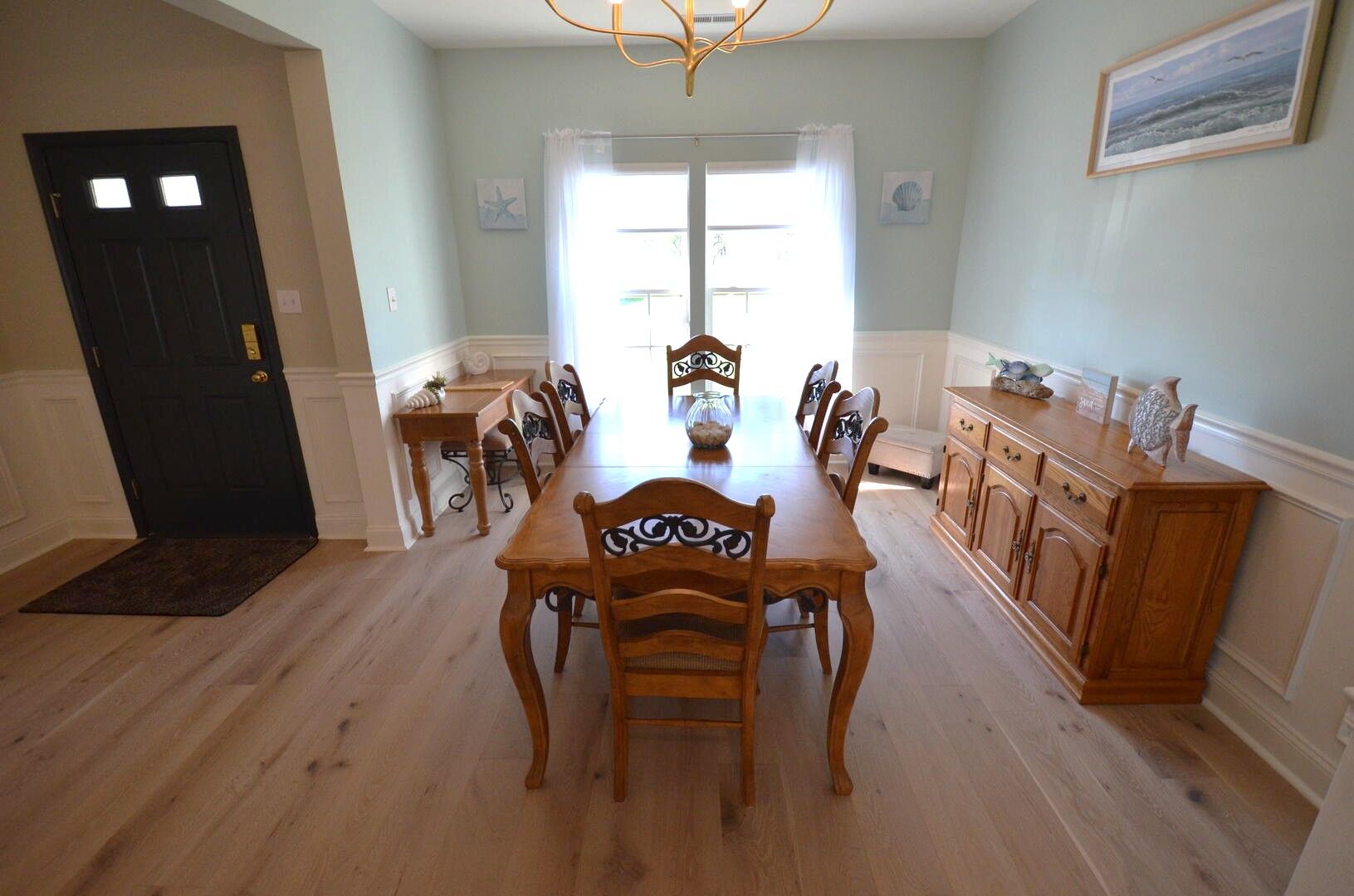 A dining room with a long wooden table, chairs, a chandelier, and a view through a window.