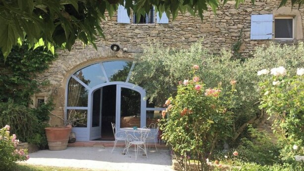 Un bâtiment en pierre doté d’une grande porte d’entrée en arc de cercle, d’une terrasse avec une table et des chaises, et d’un jardin où poussent diverses plantes et fleurs.