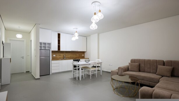 A modern kitchen with white cabinets, a wooden countertop, and a dining area with a white table and chairs. There is a brown sofa and a small round table with a gold frame in the living area. - Sanpietro Vacation Apartment 056 (Ishëm)