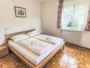 Ein ordentlich bezogenes Bett mit weißen Leinen, ein hölzernes Kopfteil, ein Nachttisch, ein gerahmtes Bild an der Wand und ein Fenster mit Vorhängen.