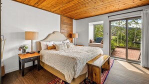 Ein Schlafzimmer mit Holzboden, einem großen Bett mit Kopfteil, zwei Nachttischen, einer Bank und Blick durch eine Schiebetür in einen Außenbereich.