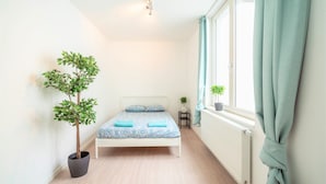 Ein Schlafzimmer mit Bett, Zimmerpflanze, kleinem Tisch und einem Fenster mit Vorhängen.