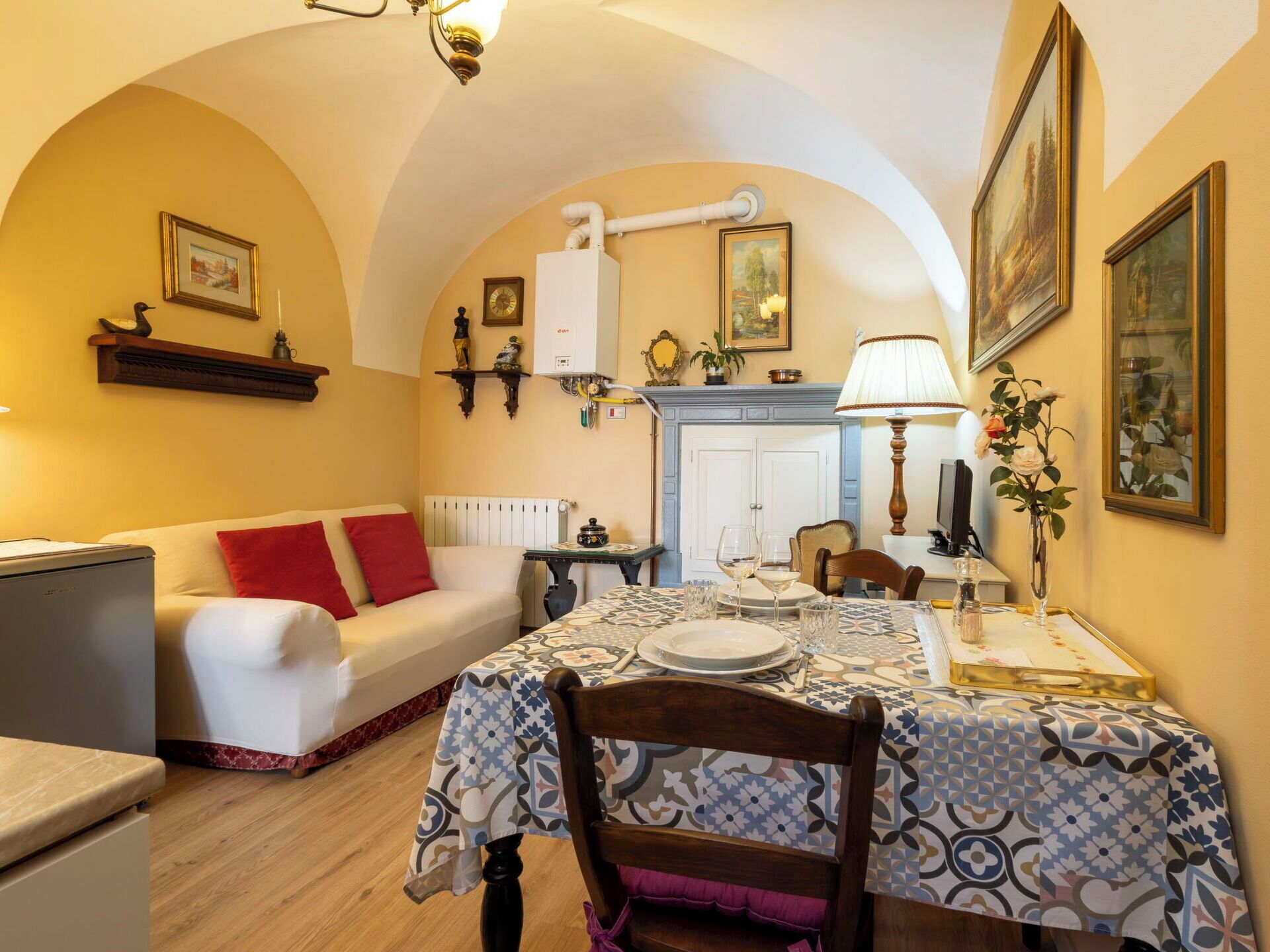A cozy dining area with a table set for two, a sofa with red cushions, a radiator, and a white cabinet.