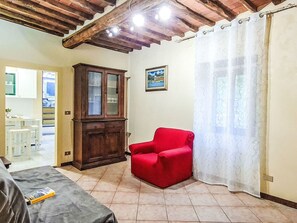 Ein Zimmer mit einem Holzkasten, einem roten Sessel, einem Bett und einem Fenster mit Vorhang.