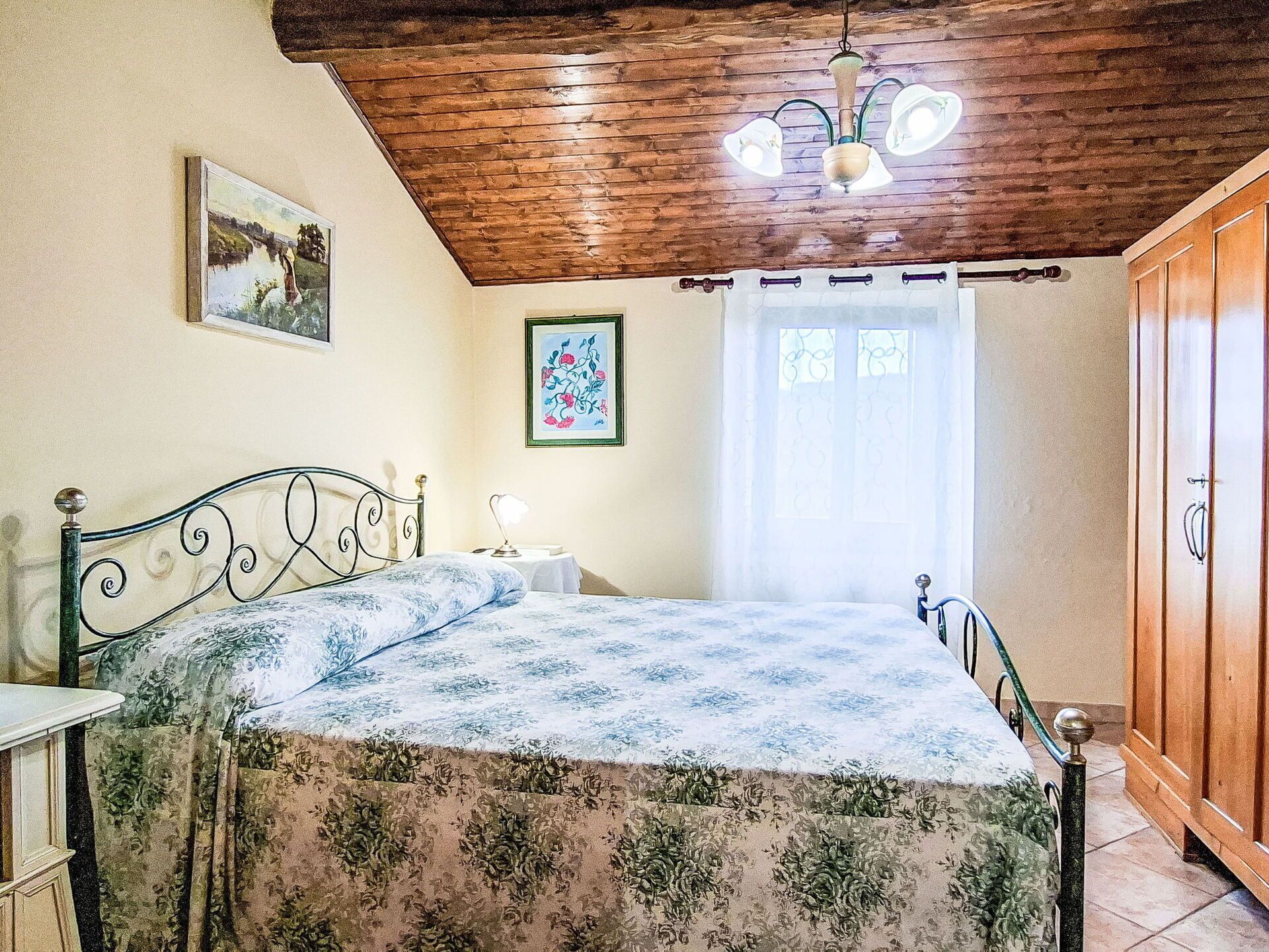 Een slaapkamer met een bed, een nachtkastje, een houten plafond en een raam met gordijnen.
