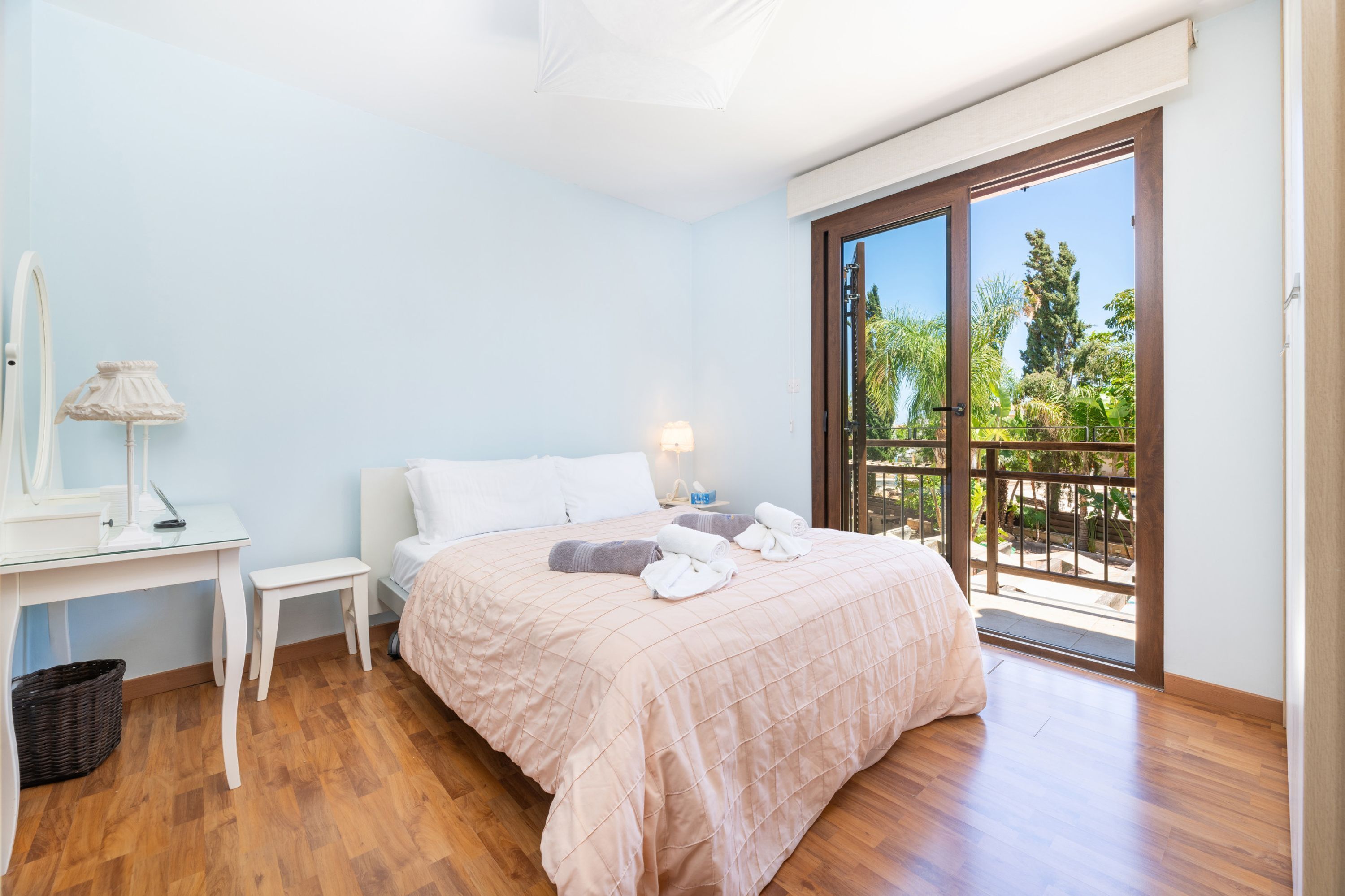 A bedroom with a bed, a desk, a lamp, a window with a view of greenery, and a door to an outdoor area.