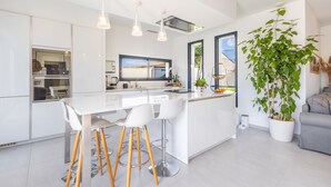 Een moderne keuken met witte kasten, een centraal eiland met krukken, hanglampen en een grote potplant.
