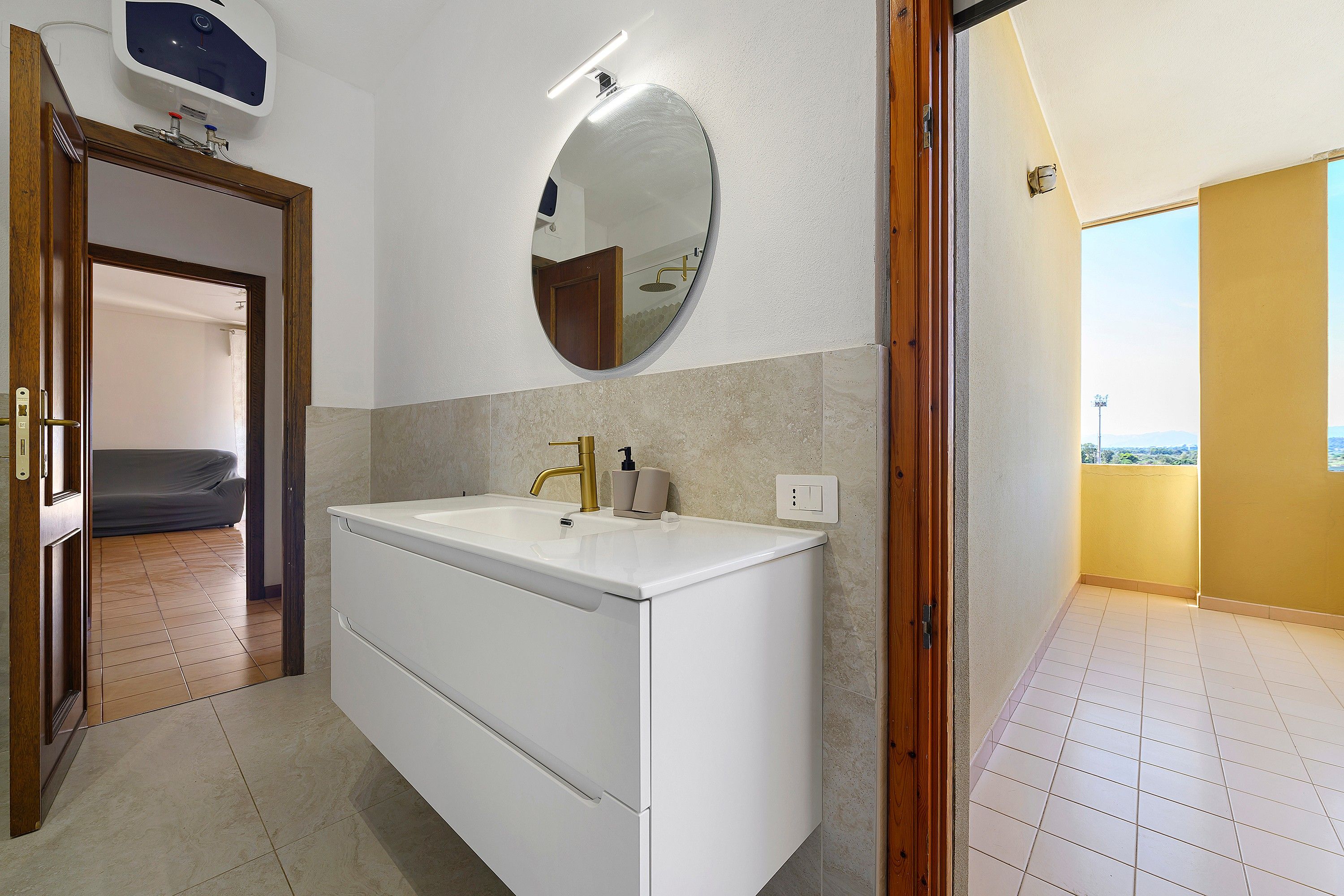 A bathroom with a white sink, gold faucet, and a round mirror. There is a door leading to another room and a view of a balcony.