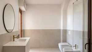 Ein modernes Badezimmer mit einem großen, ovalen Spiegel, einem weißen Waschbecken, einem Bidet und einer Duschkabine aus Glas.