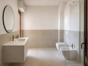 A modern bathroom with a large oval mirror, a white sink, a bidet, and a glass-enclosed shower.