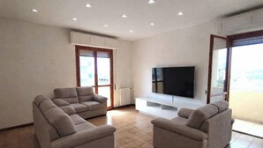Ein modernes Wohnzimmer mit einem Ecksofa, einem Flachbildfernseher und einer Schiebeglastür, die auf einen Balkon führt.