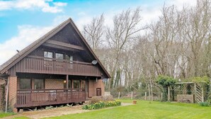 A wooden house with a balcony, surrounded by a grassy yard and trees. - Ancton Way by the Sea - sleeps 9 guests  in 4 bedrooms (Bognor Regis)