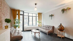 Ein möbliertes Wohnzimmer mit Backsteinwand, großen Fenstern, einer Couch, einem Couchtisch und einer Vase auf einem Sockel.